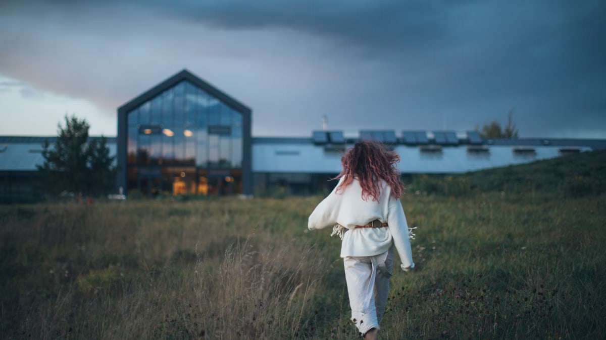 A woman walks through a grassy field towards a modern building during twilight, creating a serene and contemplative scene.